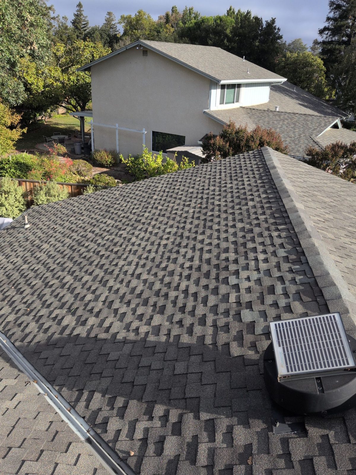 Residential roof from the ridge showing a vent, shingle field and surrounding trees